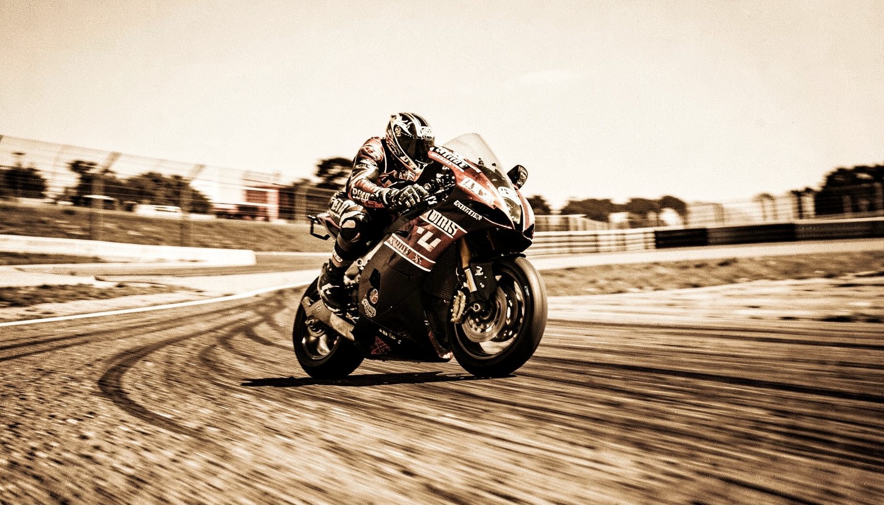 Duotone photograph of a motorcycle racer leaning into a curve on a track, warm asphalt tones against a bright sky